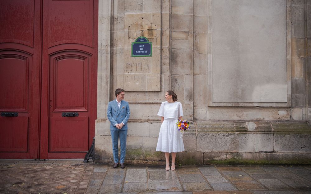 Bride and groom stand apart by a red door on a Paris street, bride holding colorful bouquet.