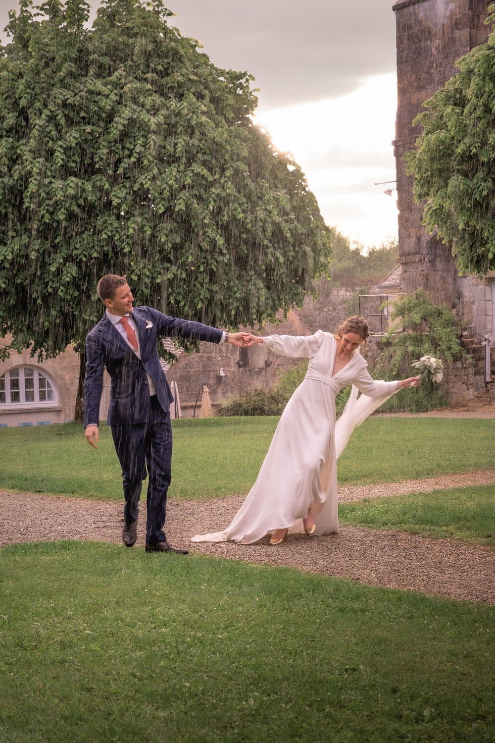 Bride and groom dancing joyfully in the rain on their wedding day in a lush garden setting.