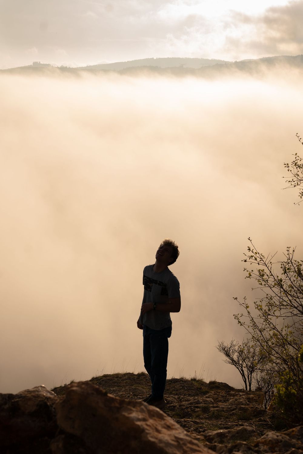 Person standing on rocky cliff edge overlooking a misty valley with fog-filled mountains at golden hour.