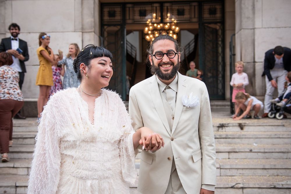 Joyful newlywed couple laughing on stone steps after wedding ceremony, bride in white feathered dress, groom in tan suit.