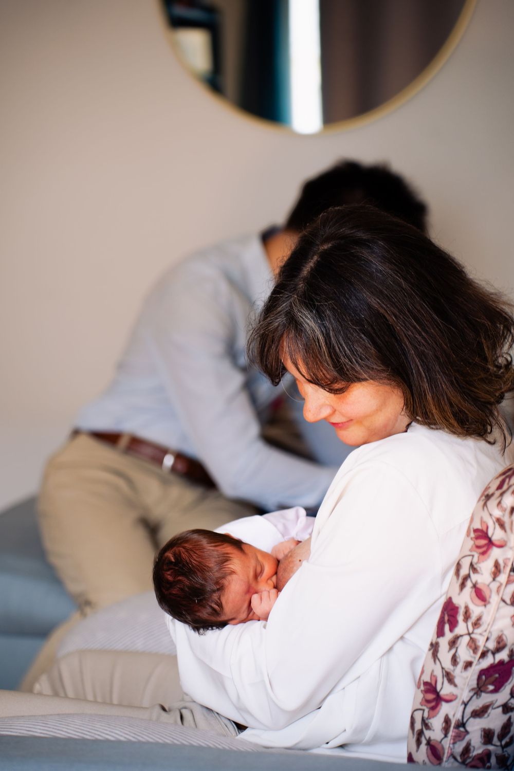 A mother tenderly holds her newborn baby while sitting indoors, with a man visible in the background.
