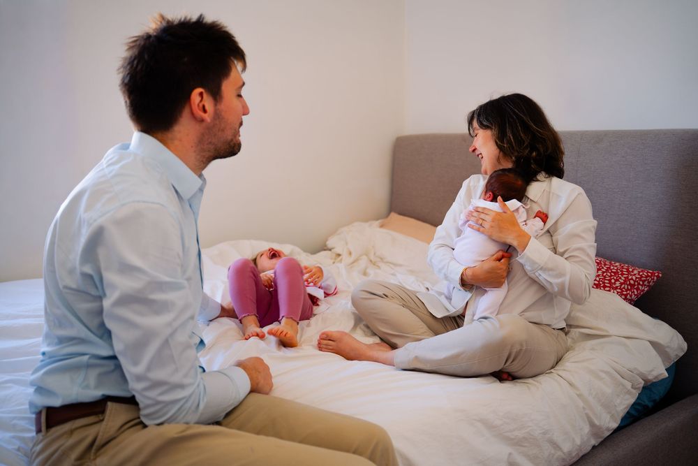Pregnant woman sitting on bed holding stuffed animal while man sits beside her in bright bedroom.