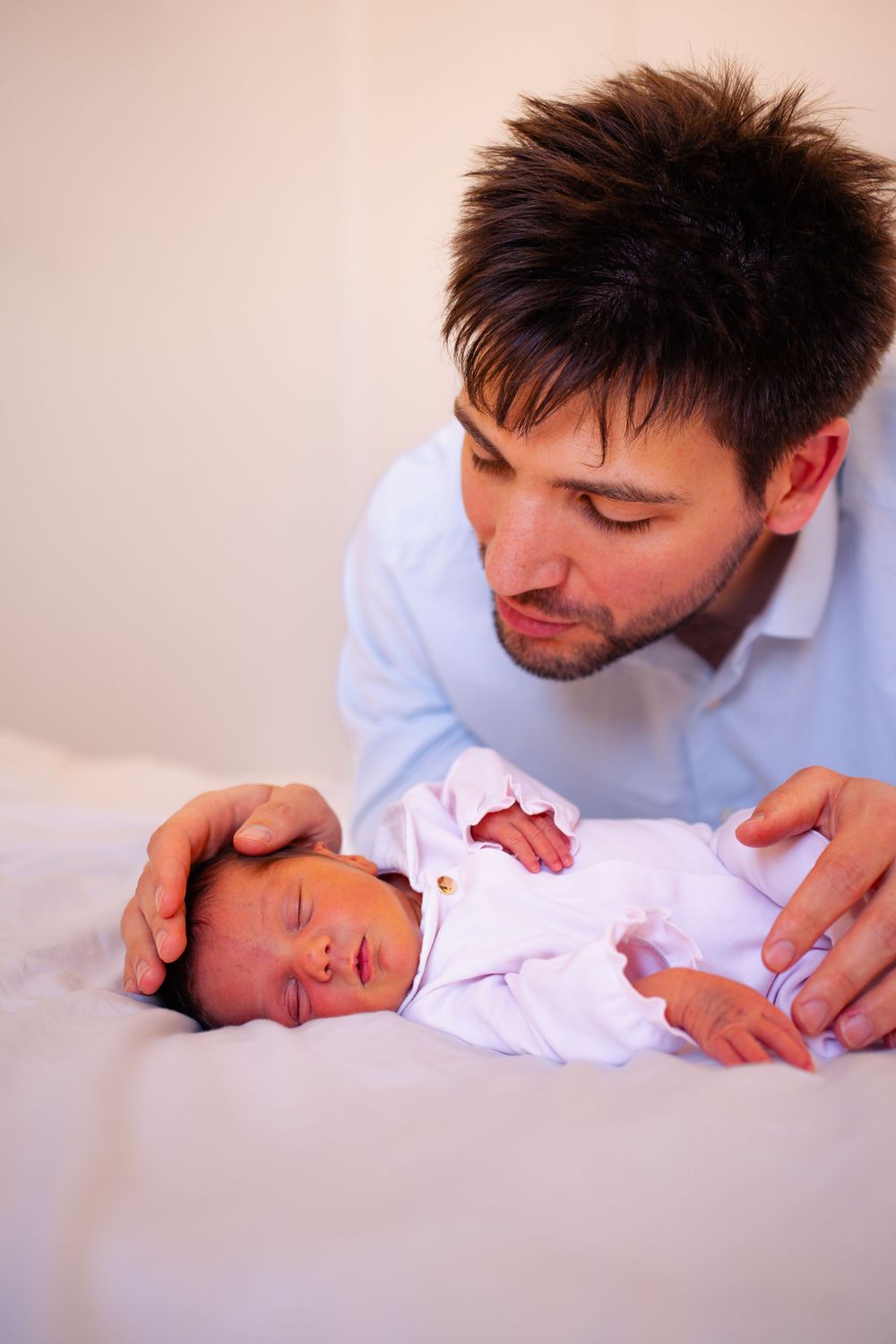 A young father in a blue shirt gently cradles his sleeping newborn baby on a white bed.