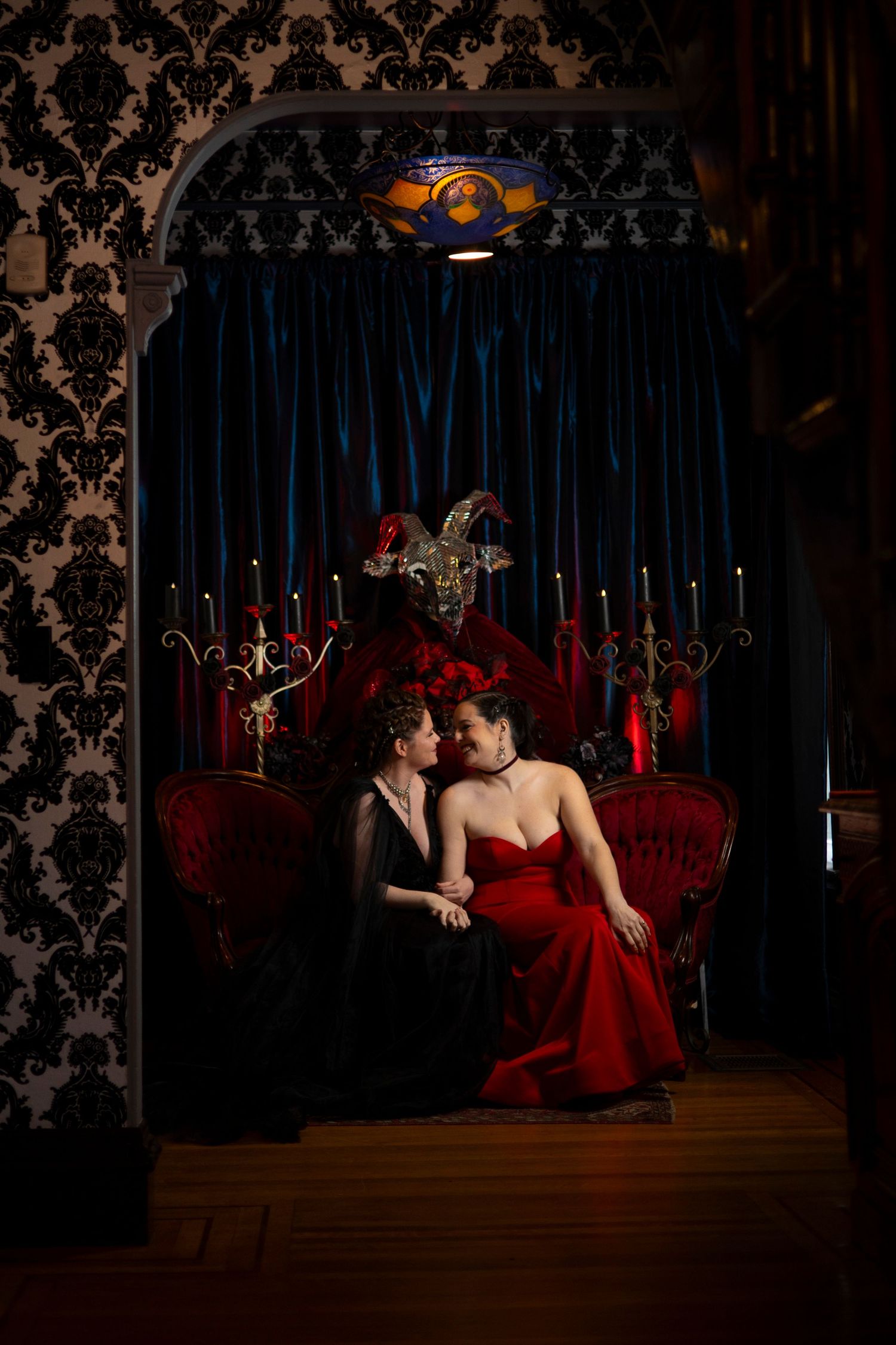 Romantic couple in formal attire sitting on red velvet couch against dark damask wallpaper and blue curtains.