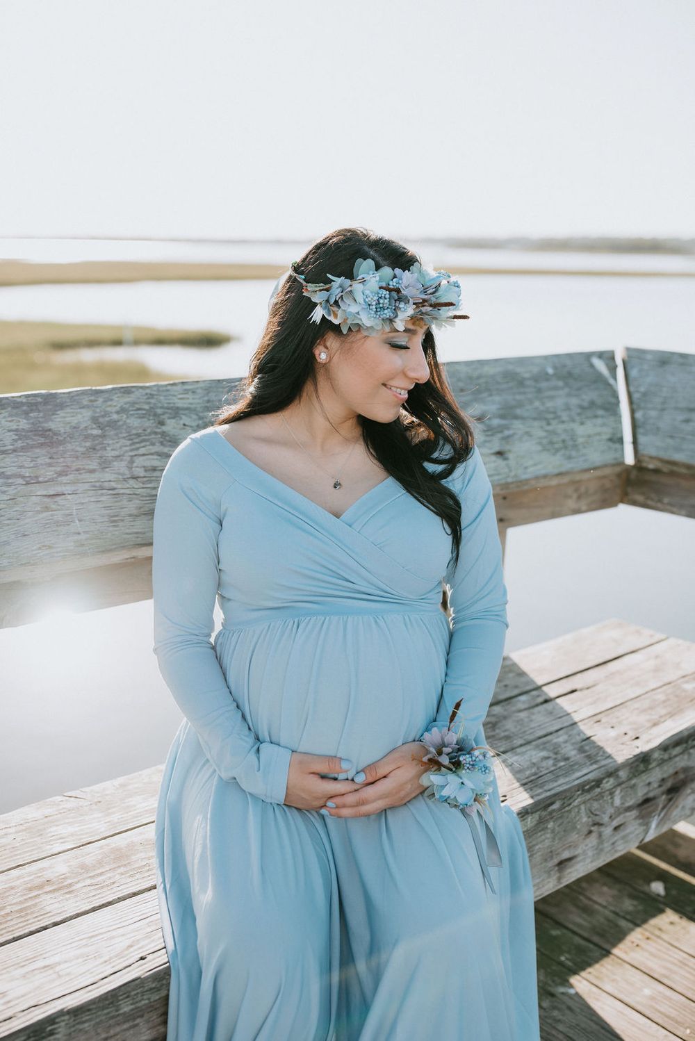Expectant woman in flowing light blue dress and floral crown stands on wooden dock by waterfront in soft sunlight.