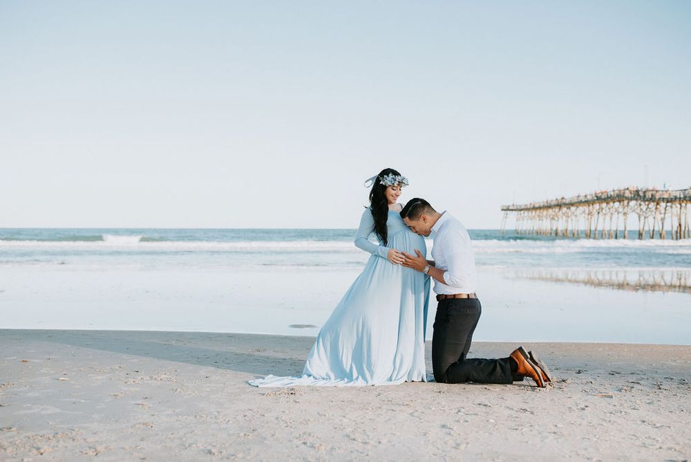 A couple in light blue outfits walks along a serene beach with a wooden pier in the background.