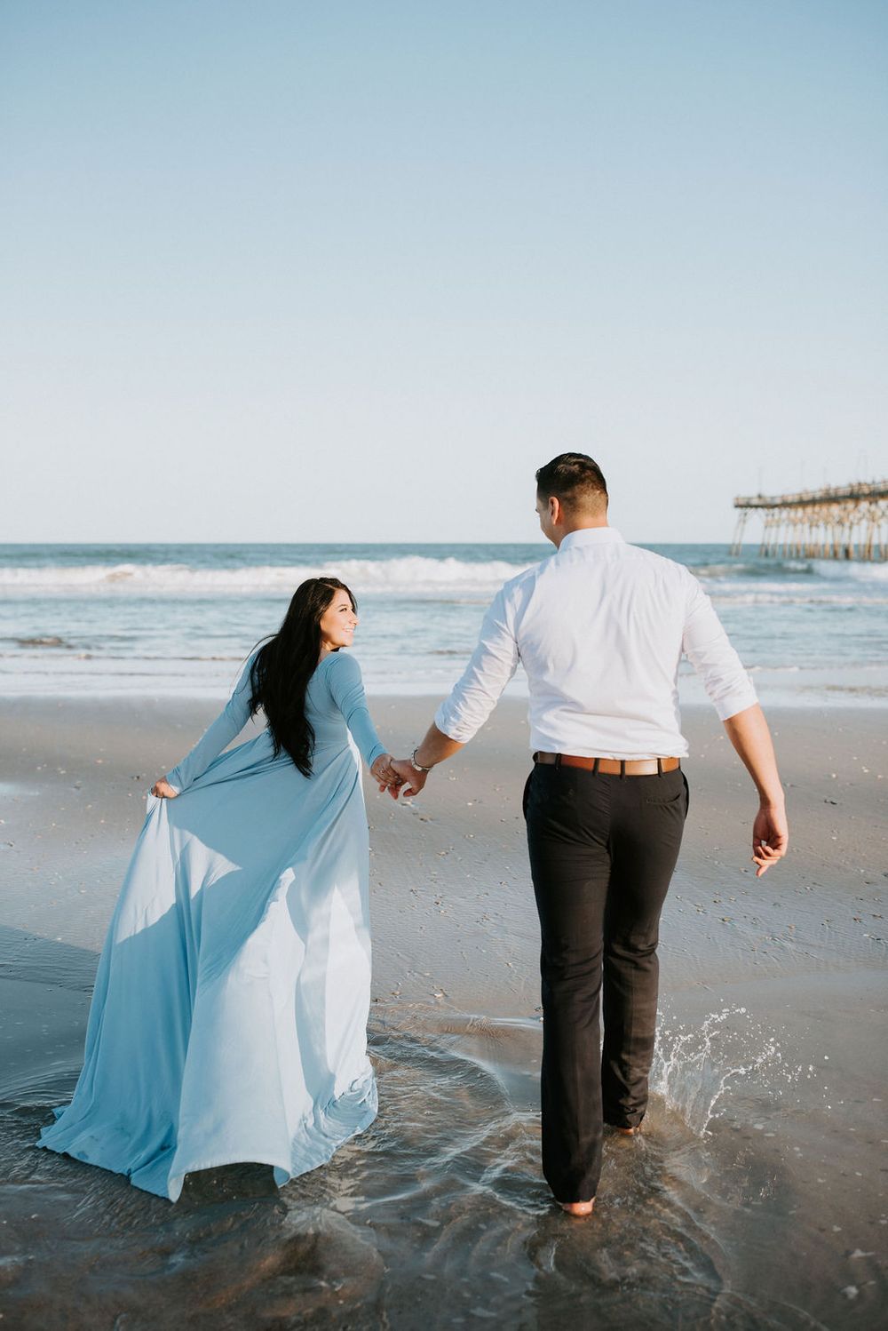 A couple walks hand in hand on a beach at sunset, with the woman wearing a flowing light blue dress near ocean waves.