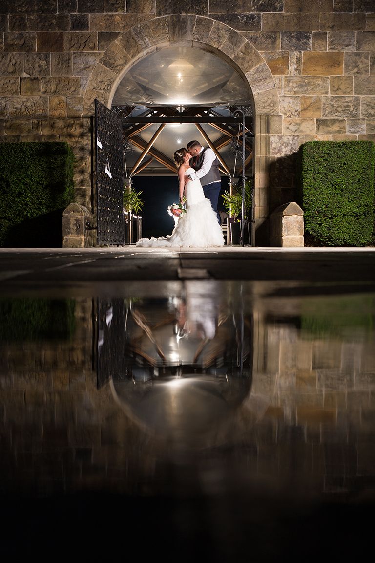 A romantic wedding couple embraces in front of a stone wall with their reflection visible in a dark pond below.