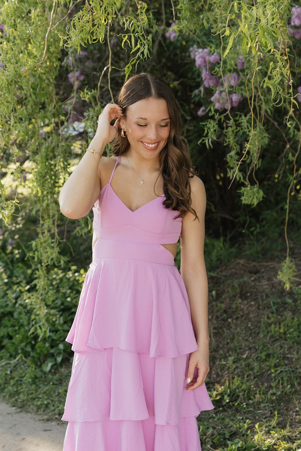 A person in a flowing pink tiered dress poses outdoors against lush green foliage in a garden setting.