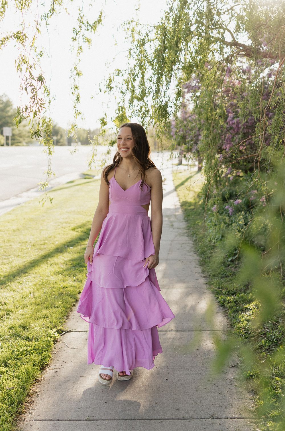 A flowing lilac dress twirls gracefully on a sunlit garden path bordered by lush greenery and blooming flowers.