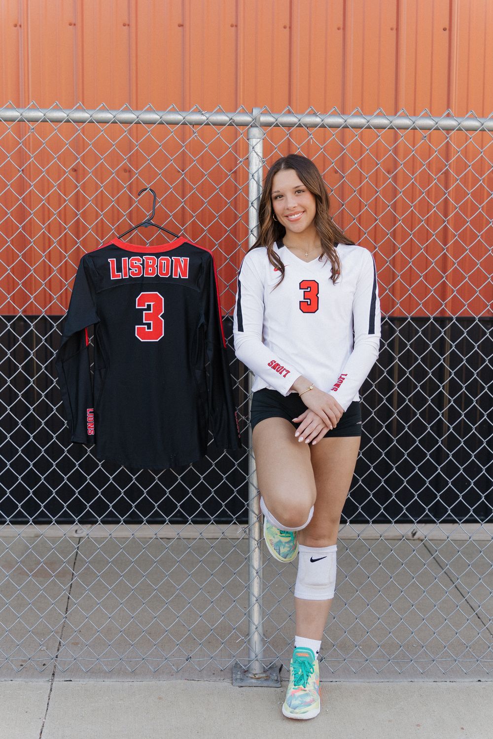 Athletic volleyball player in white jersey with number 3 poses against chain link fence with red background.