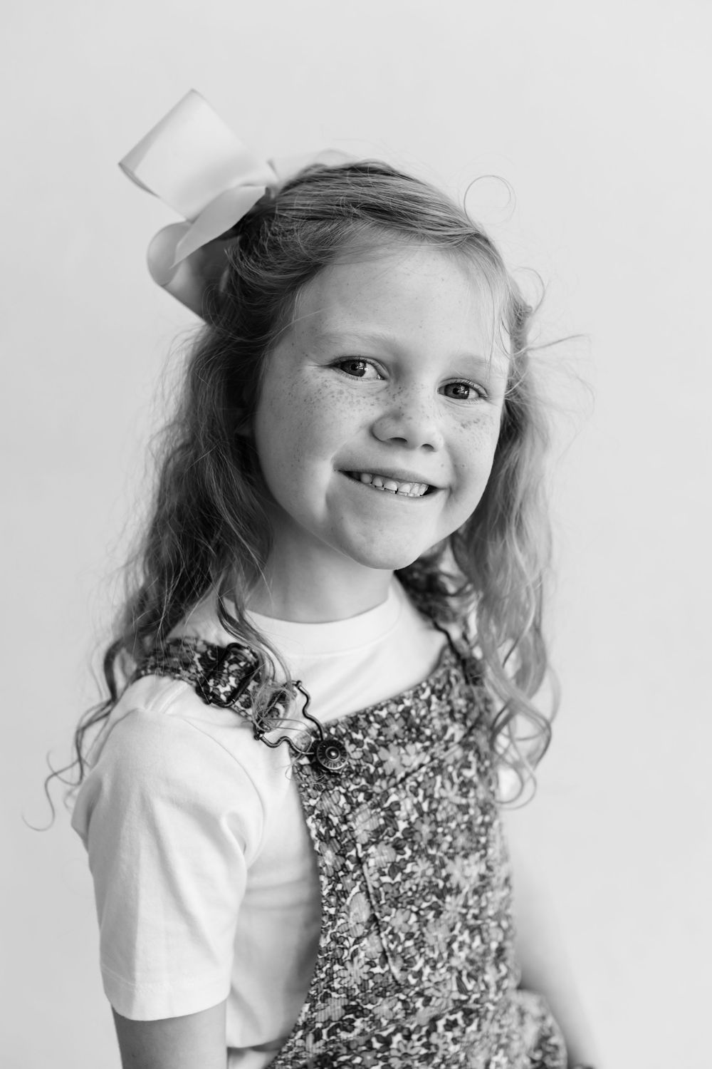 Black and white portrait of a smiling young child wearing a white shirt and sparkly dress with a bow in their hair.