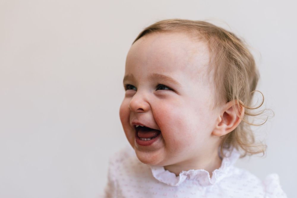 A laughing baby with blonde curls wearing a white outfit expresses pure joy in a series of candid photos.