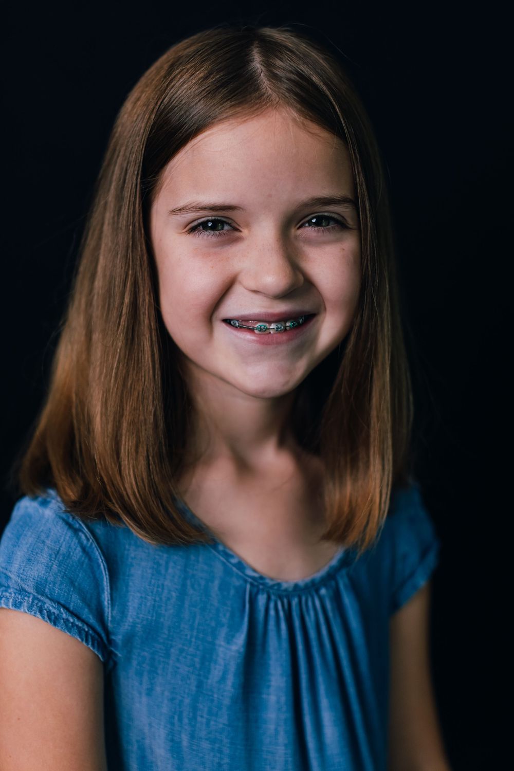 Portrait of a cheerful young person with straight brown hair wearing a blue top against a dark background.