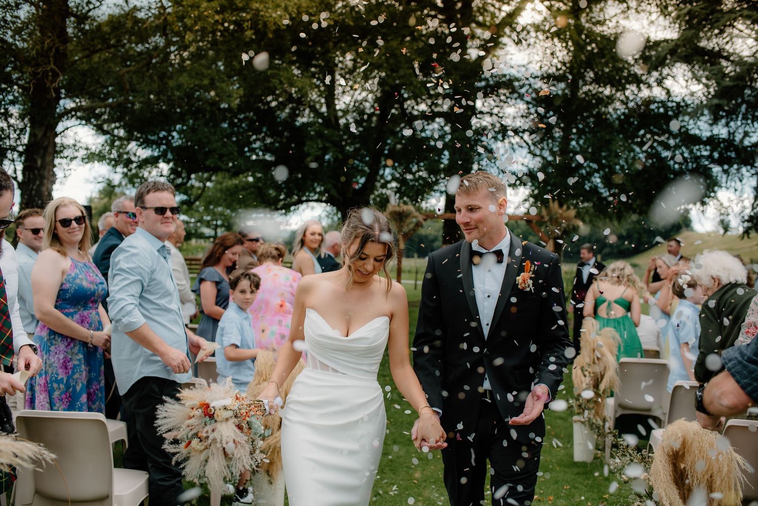 Wedding guests gather outdoors at a summer reception with trees and string lights creating a festive atmosphere.