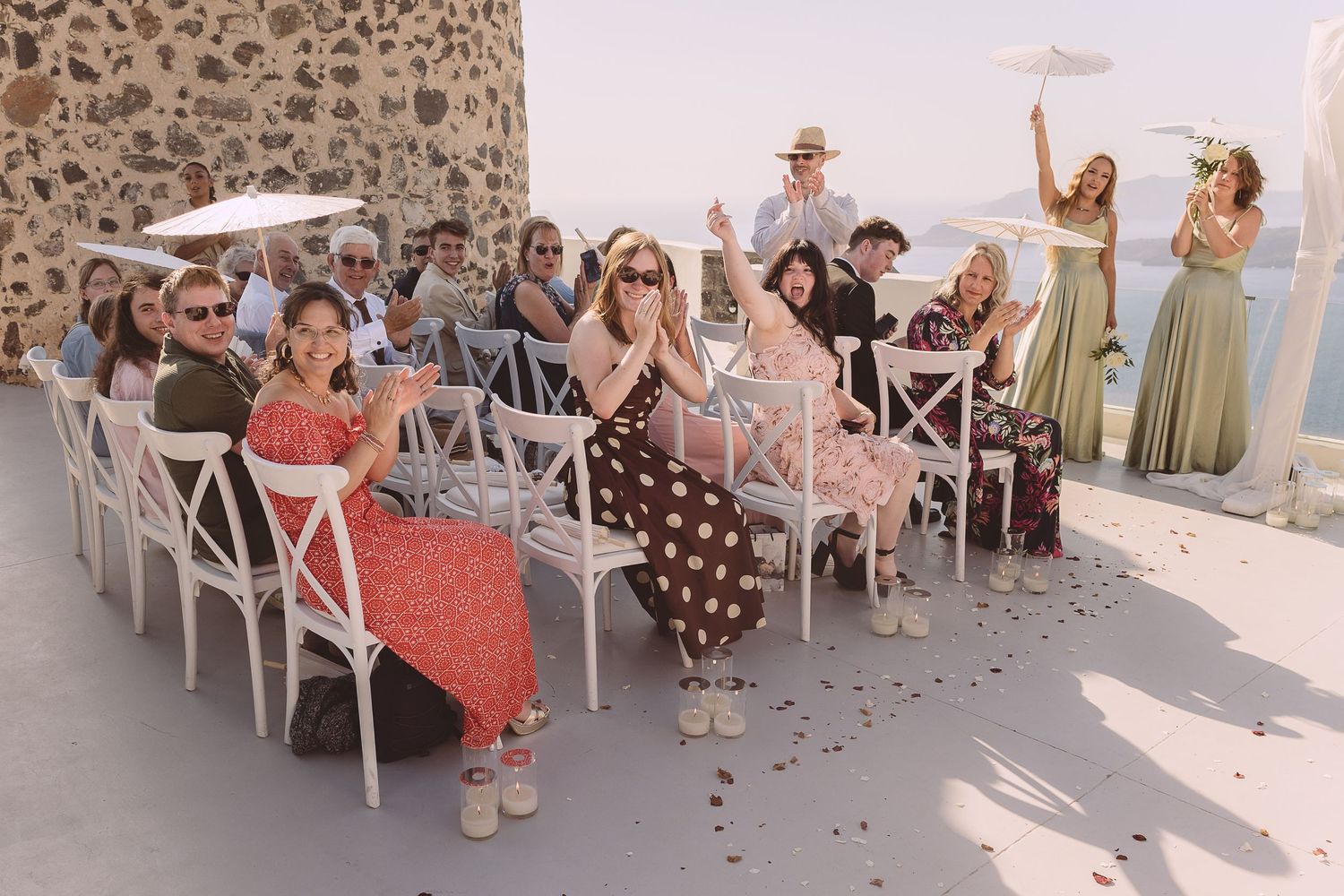 Wedding guests seated in white crossback chairs react joyfully during an outdoor destination wedding ceremony with stone wall backdrop.