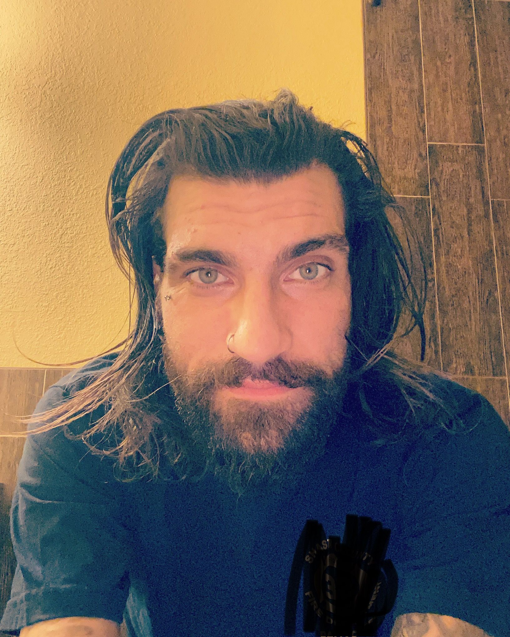 A person with long dark hair and a full beard wearing a black shirt poses against a yellow wall.
