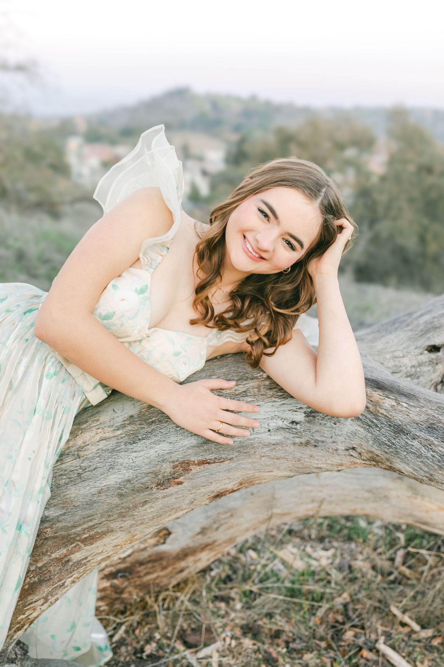 A person in a flowing white dress lays gracefully across fallen tree branches in a dreamy outdoor photoshoot.