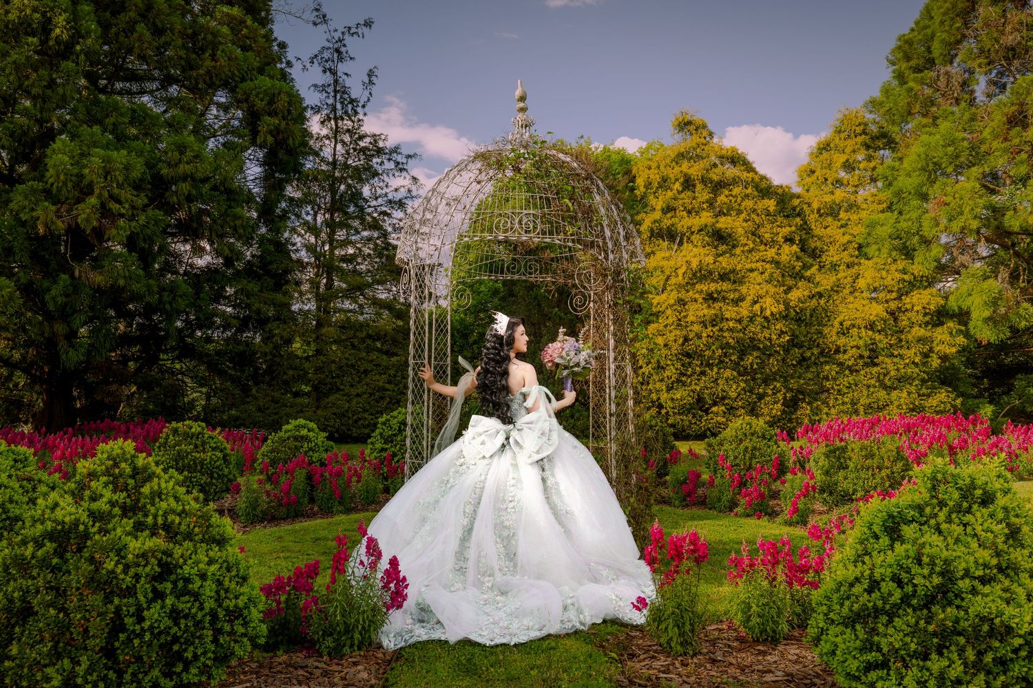 A romantic garden wedding scene with an ornate metal gazebo surrounded by blooming flowers and lush greenery at sunset.