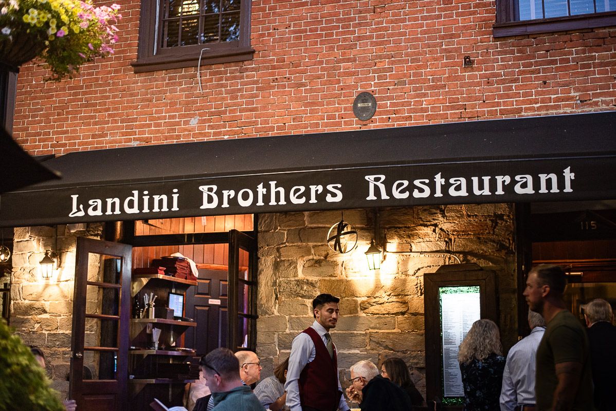 A couple shares a romantic moment outside Landini Brothers Restaurant with brick walls and flowers in the background.