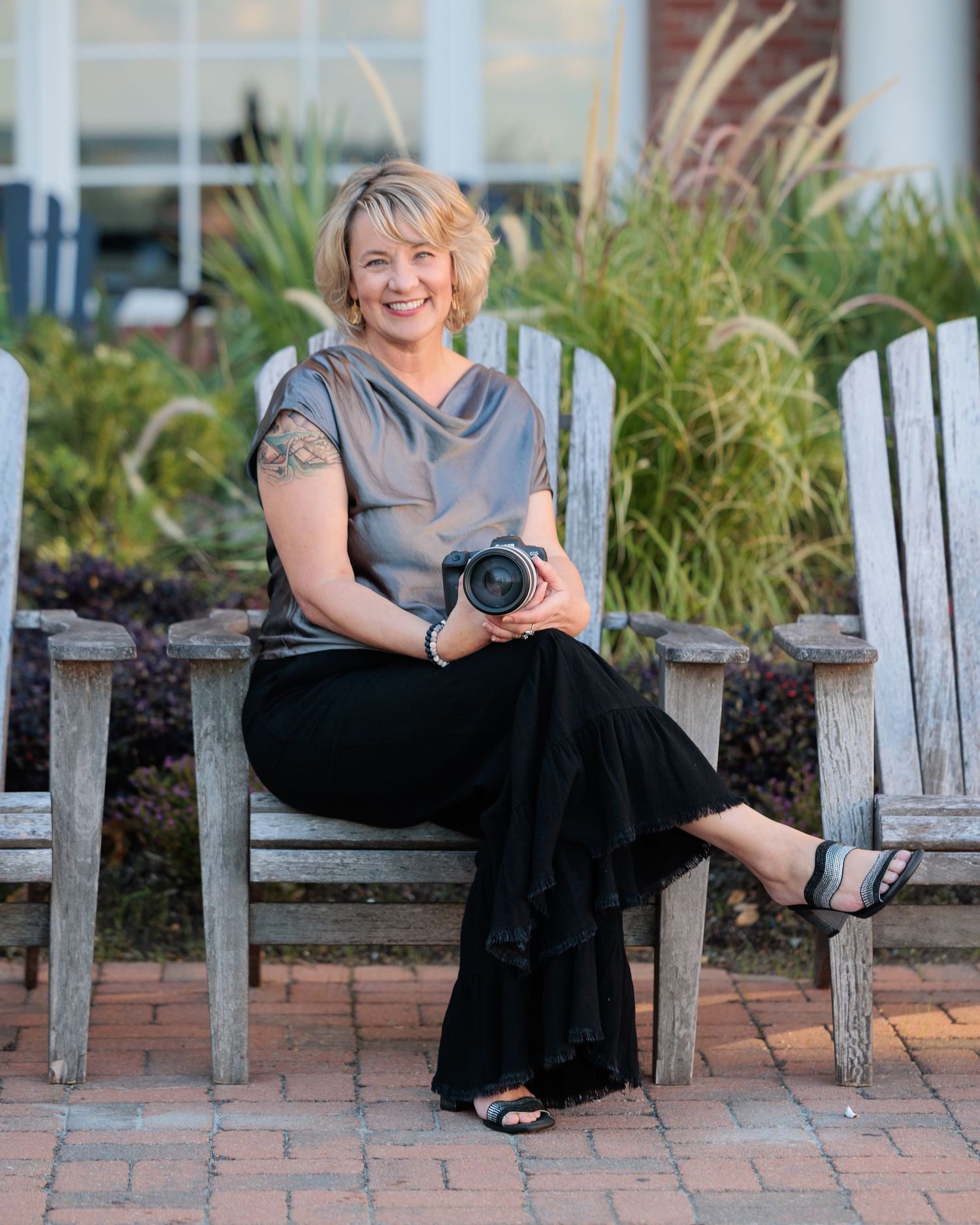 A photographer sits on a wooden Adirondack chair on a brick patio with beach grass in the background.