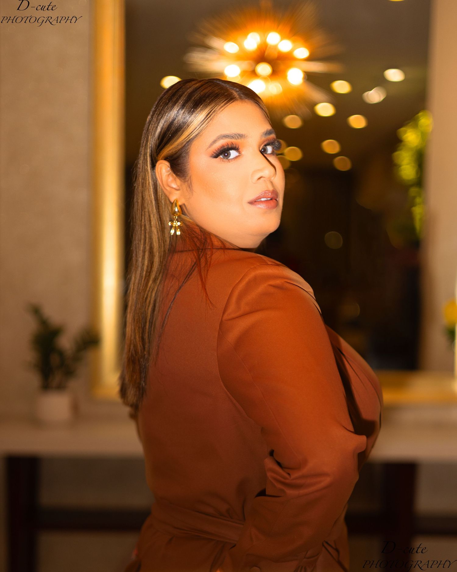 Woman in brown blazer with highlighted hair posing over shoulder in bokeh light setting.