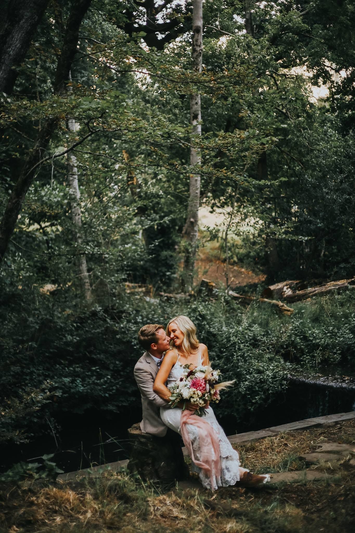 A bride and groom share a romantic moment seated by a forest creek, bride holding a colorful bouquet in a flowy gown.
