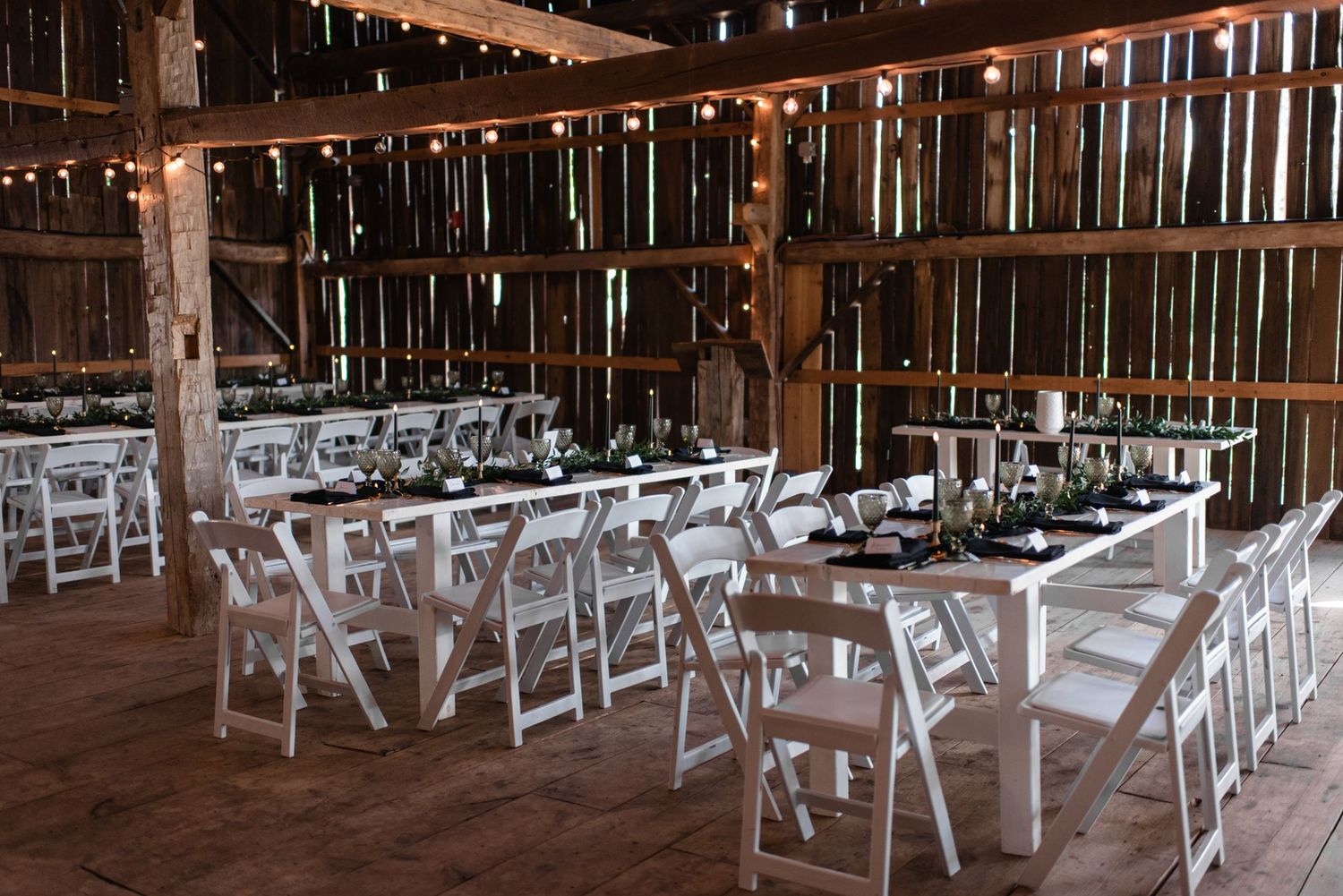 White folding chairs are arranged in rows inside a rustic wooden barn venue with string lights overhead.
