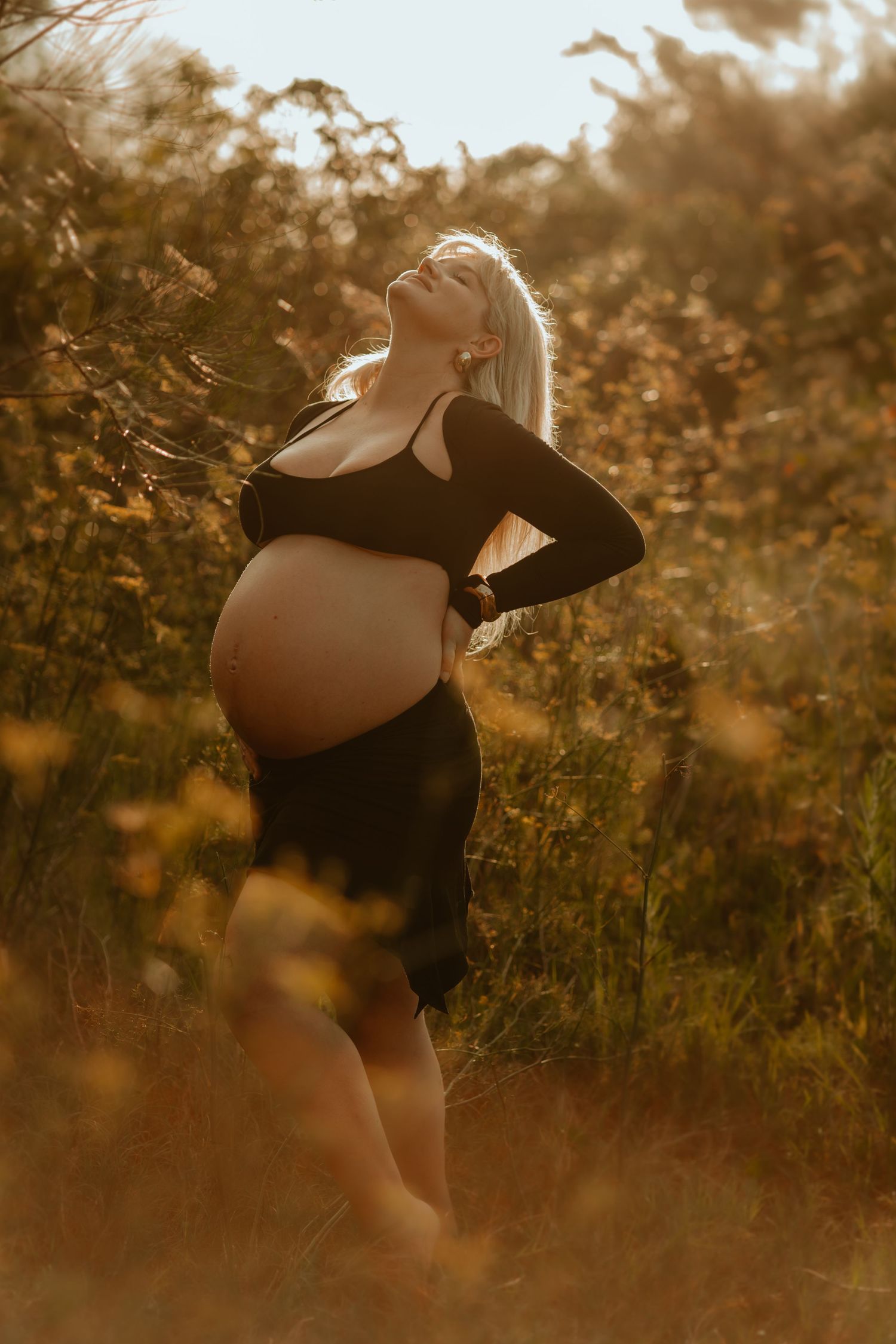 A silhouette of a pregnant woman in black dress poses gracefully in a golden sunset field with wildflowers at Kurnell Beach Bonna Point