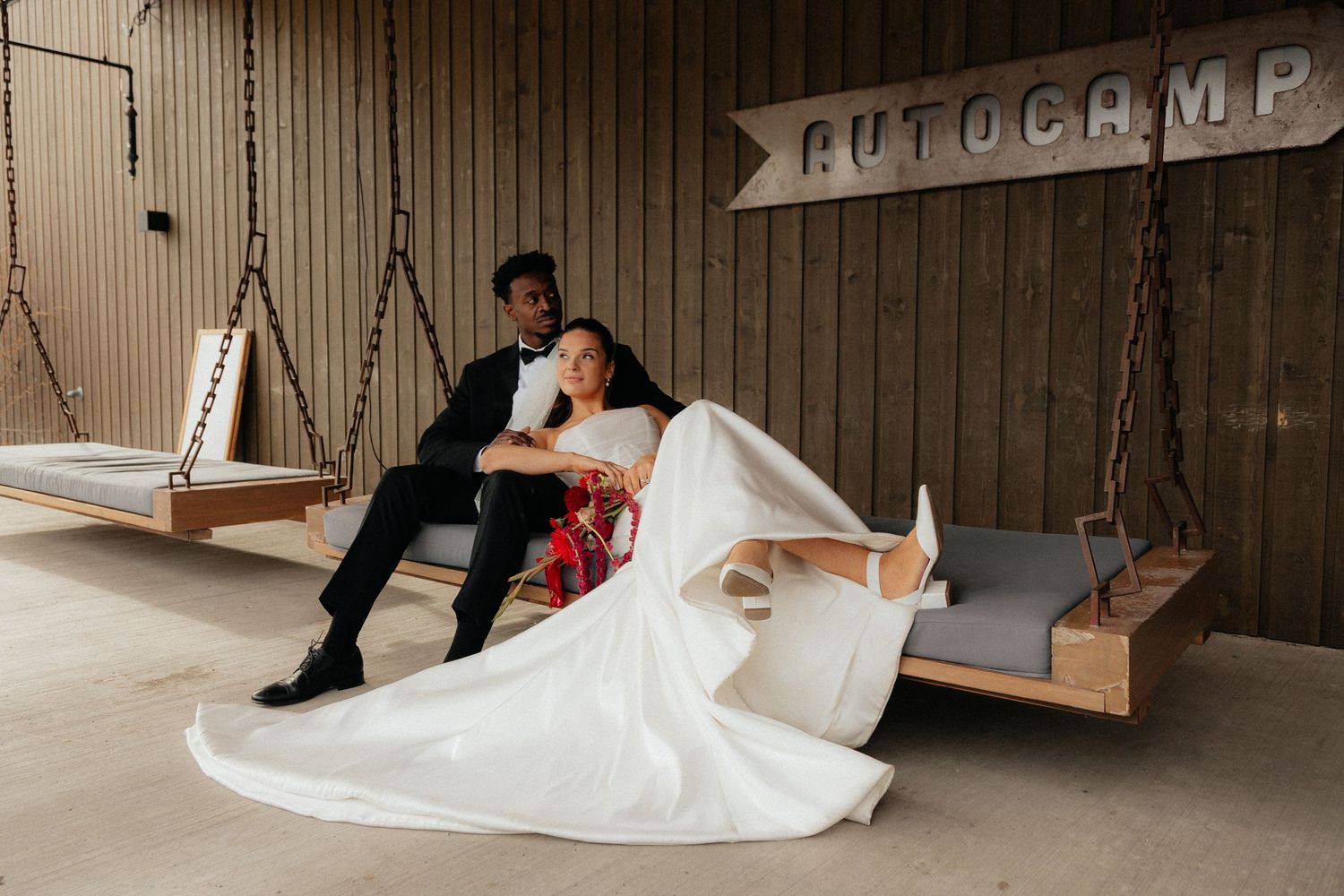 Bride and groom relaxing on hanging wooden swings inside a rustic AutoCamp venue.