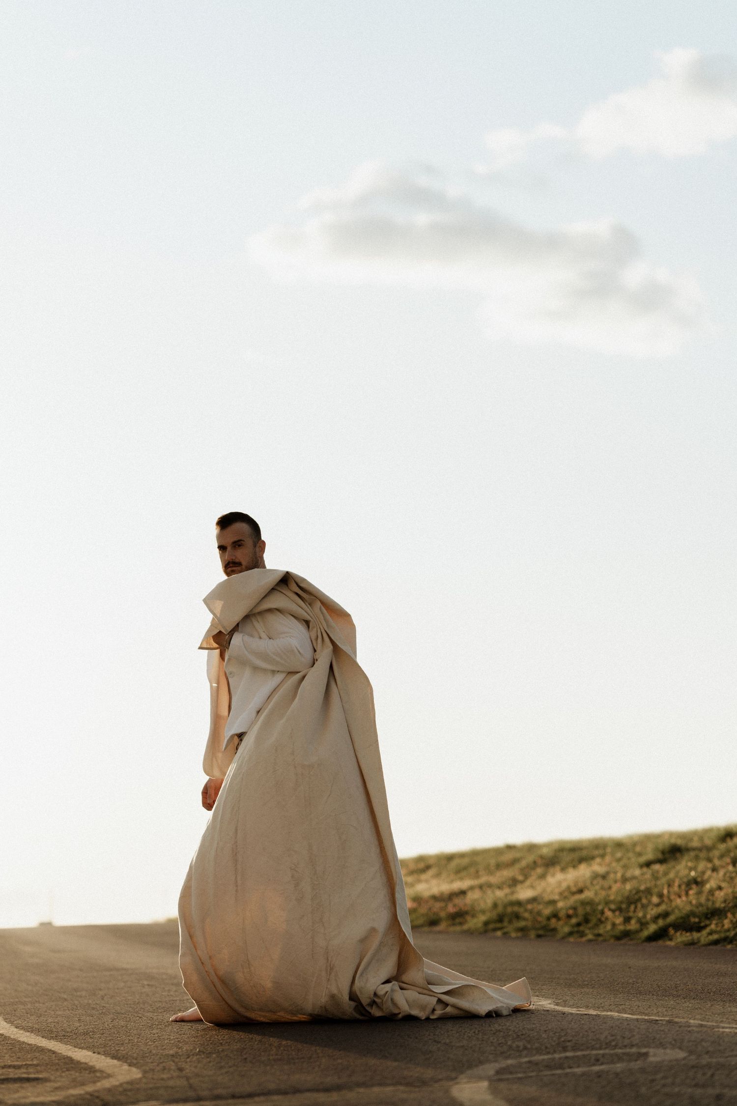 A man draped in a flowing cream-colored fabric stands on a road under a pale sky.