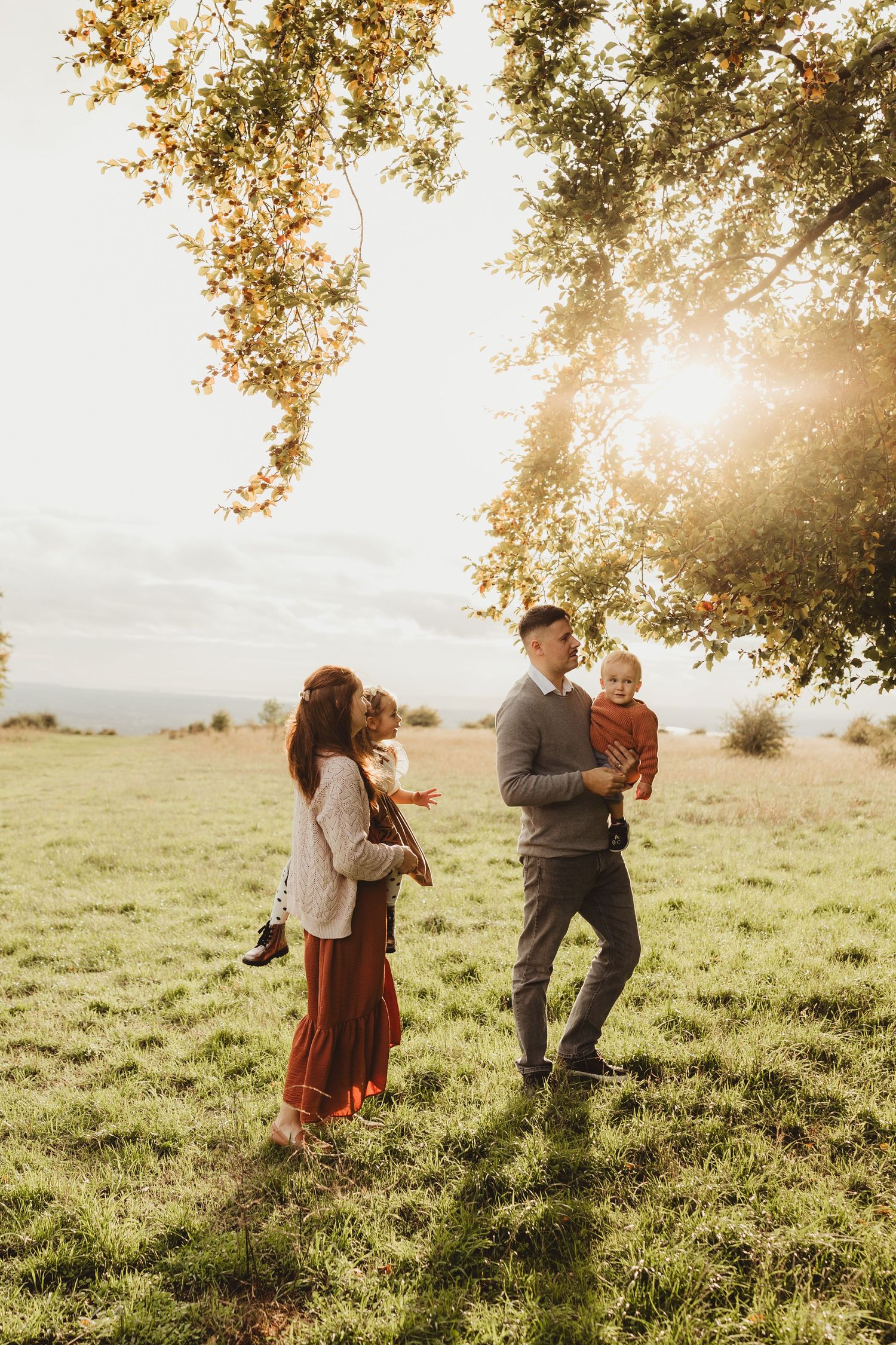 Family enjoying golden sunset in grassy meadow with sunlight streaming through autumn tree branches.