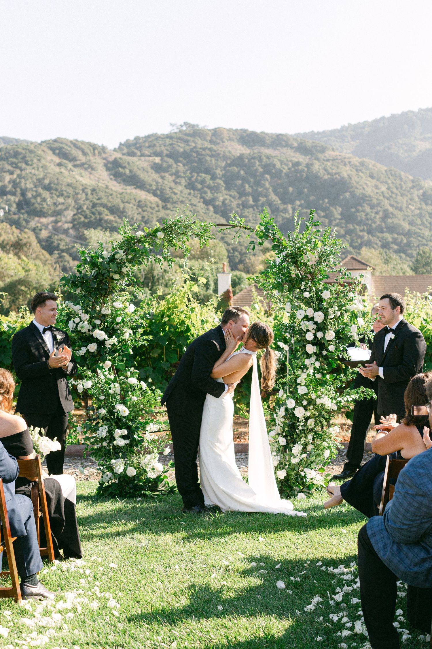 Couple embraces during outdoor garden wedding ceremony under a lush floral arch with white roses and greenery.