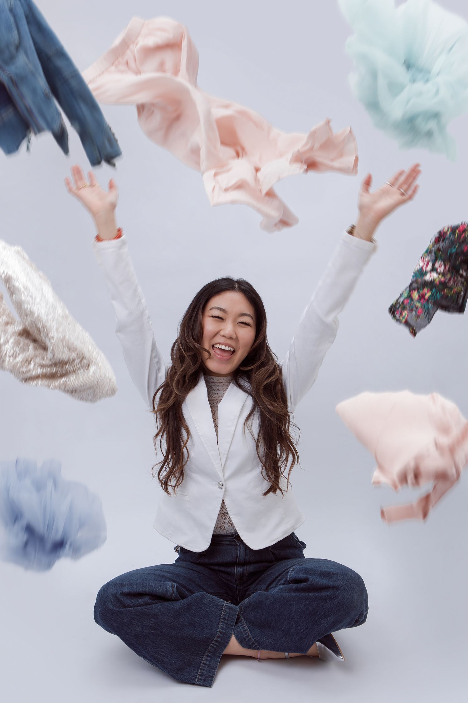 A joyful person in a white blazer and jeans sits cross-legged while colorful fabrics float through the air around them.