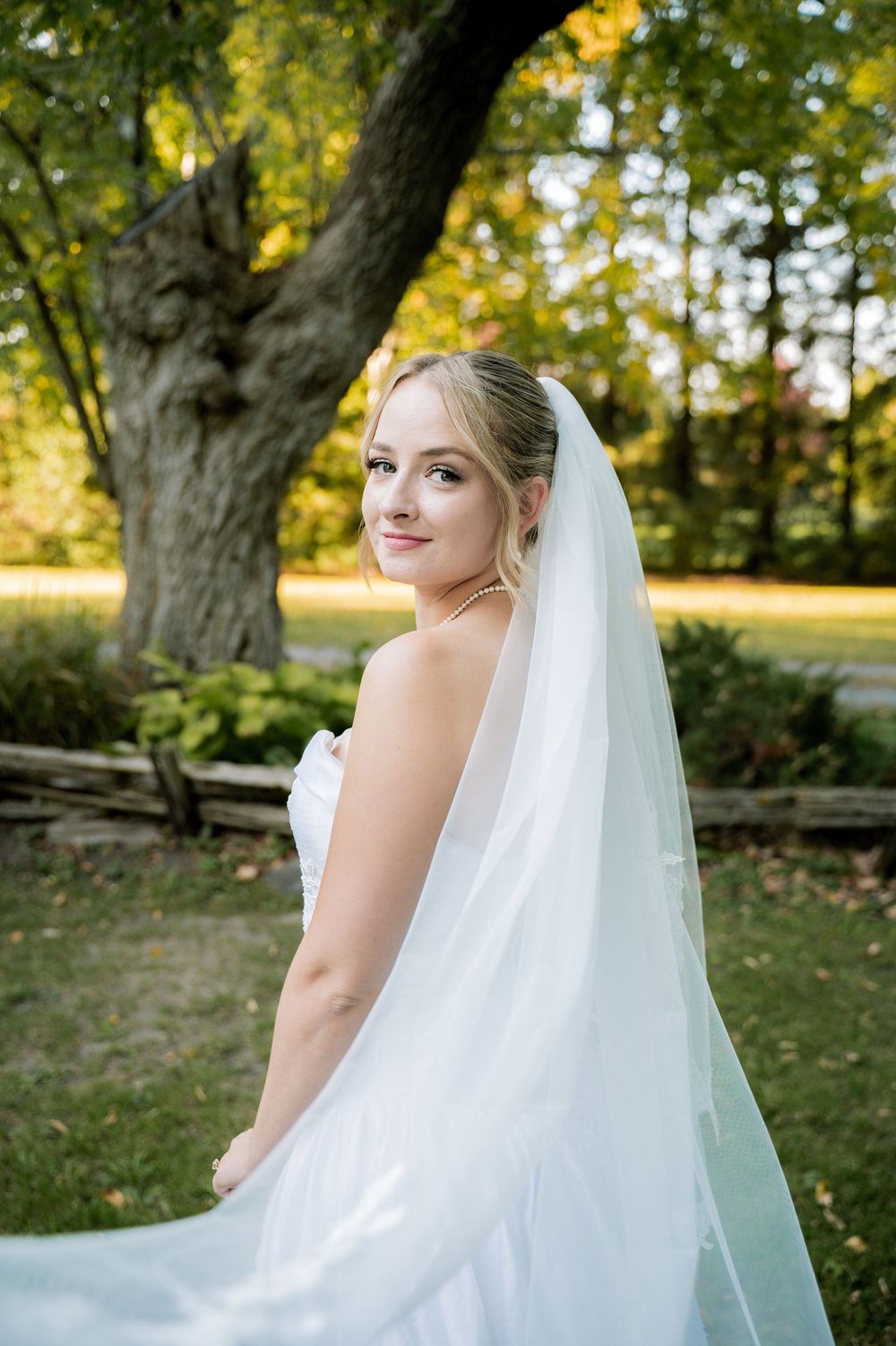 A bride in a white wedding dress stands in a garden setting with soft autumn light filtering through the trees.