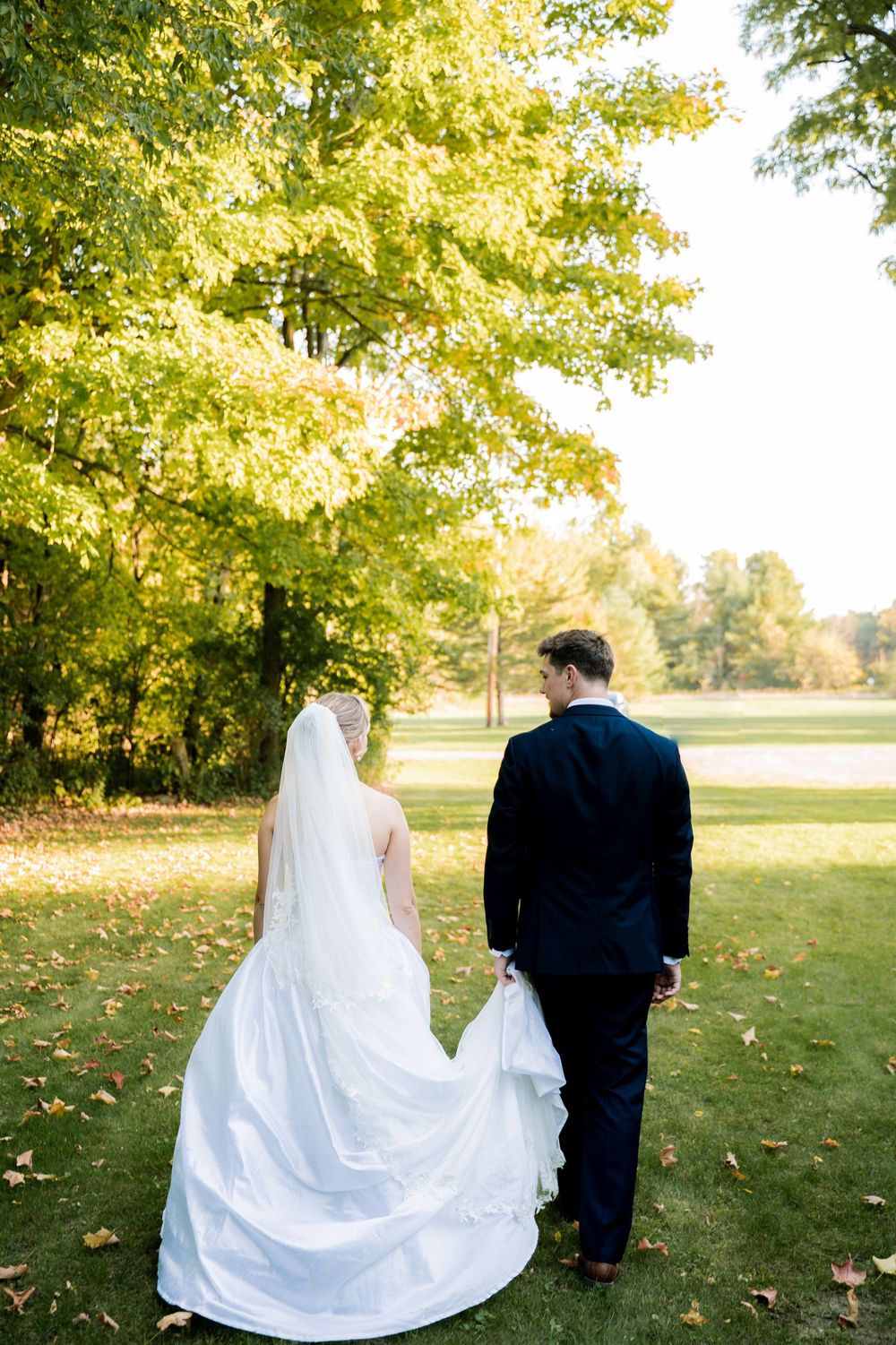 A couple in wedding attire walks together on grass with autumn trees in the background on a sunny day.