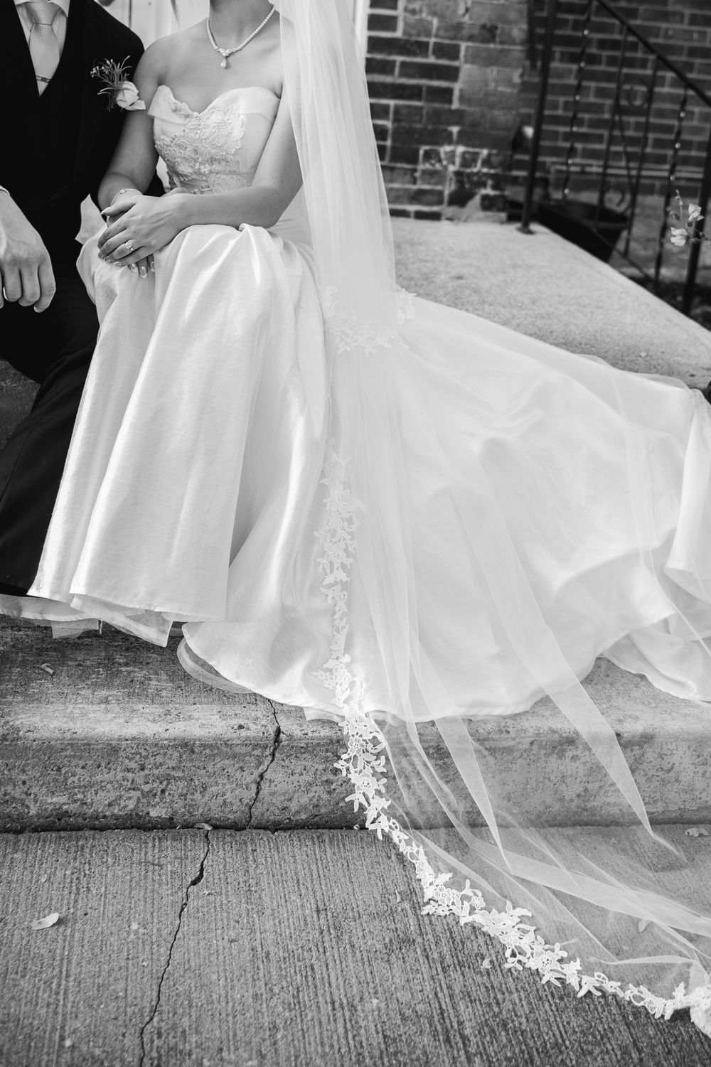 A close-up black and white detail of a flowing wedding dress with delicate lace veil trailing on concrete steps.