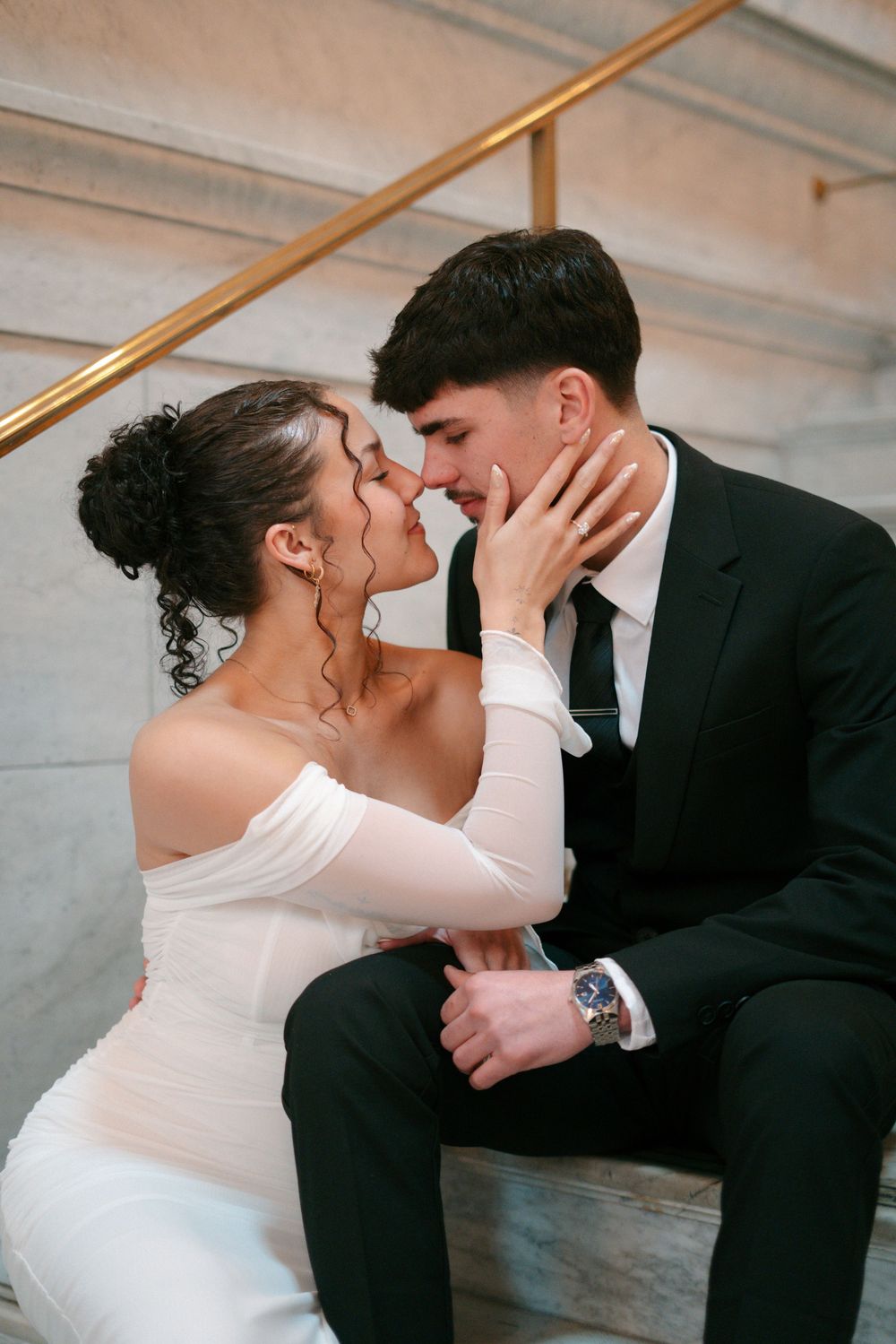 A bride in a white off-shoulder gown and groom in a black suit share a romantic moment on marble steps.