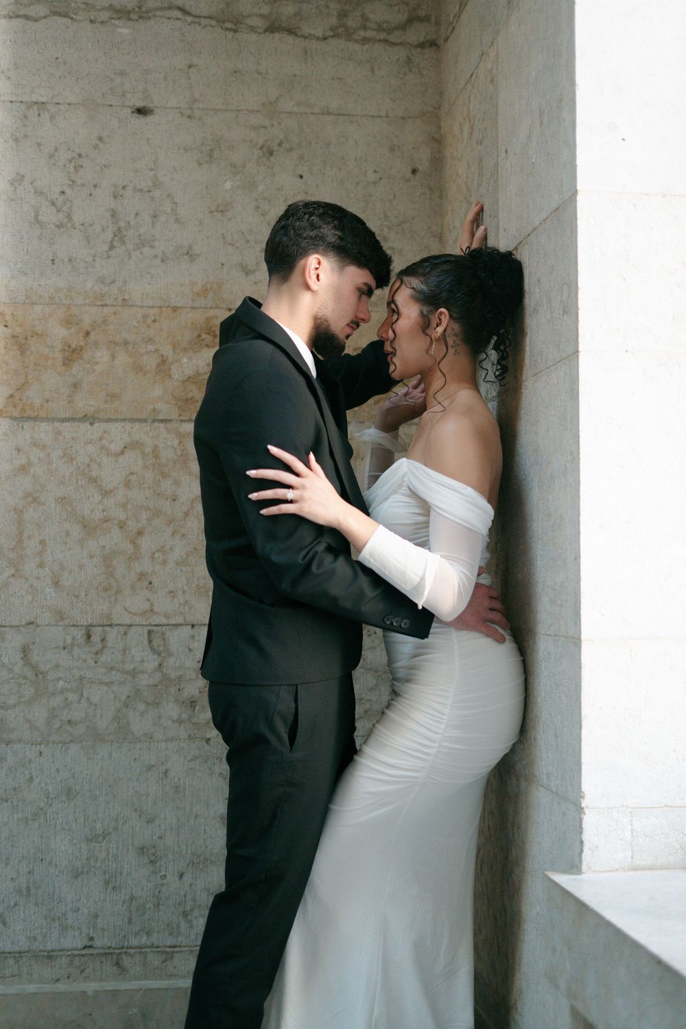 Romantic couple in wedding attire embracing against marble wall, bride in white off-shoulder gown, groom in black suit.