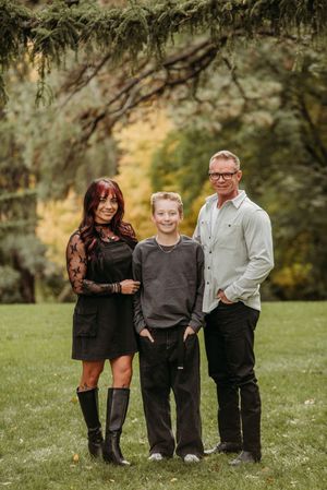 A family poses together outdoors on grass with trees in the background during a fall photo session.
