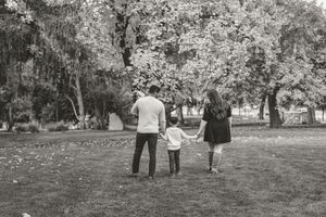 A family walks together holding hands in a park with trees and fallen leaves in black and white.