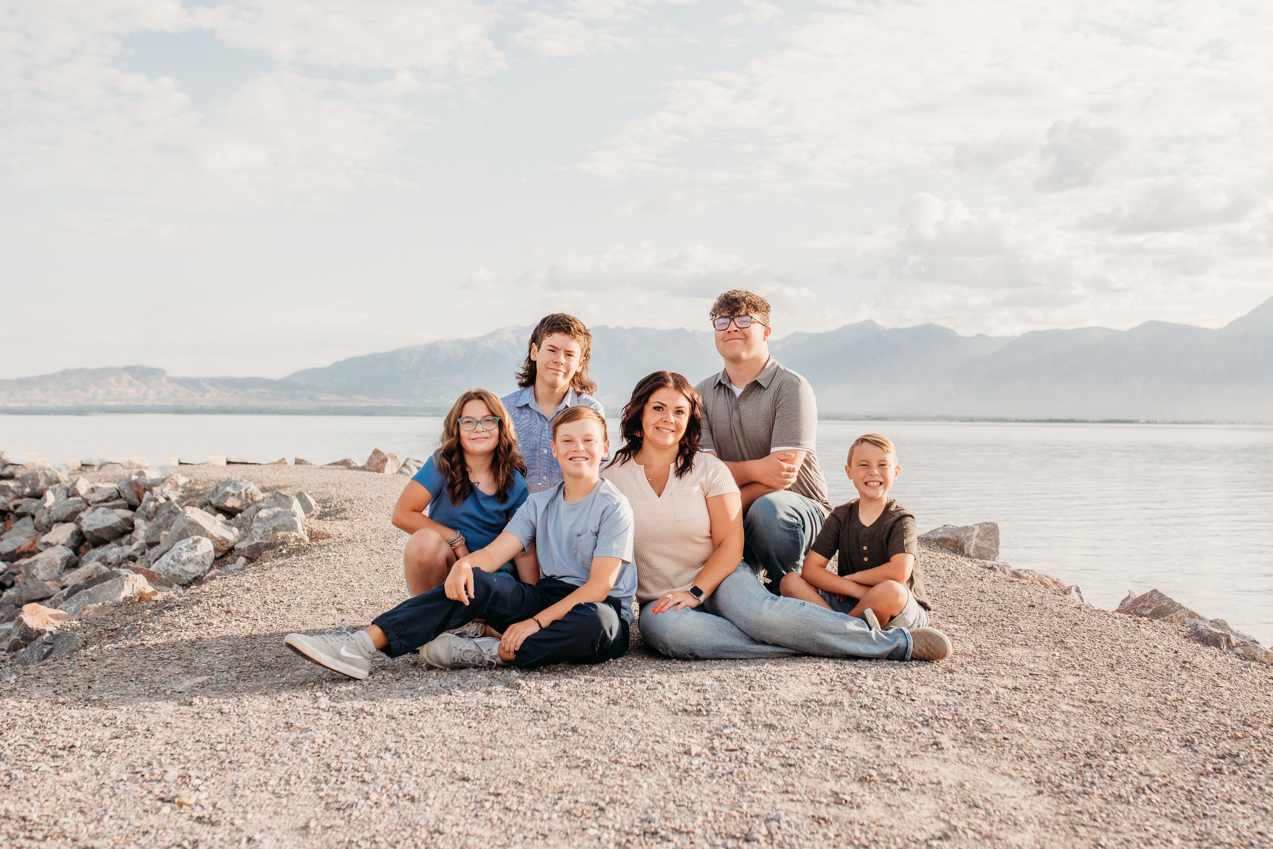 A family relaxes together on a rocky beach shore with mountains and water in the background.
