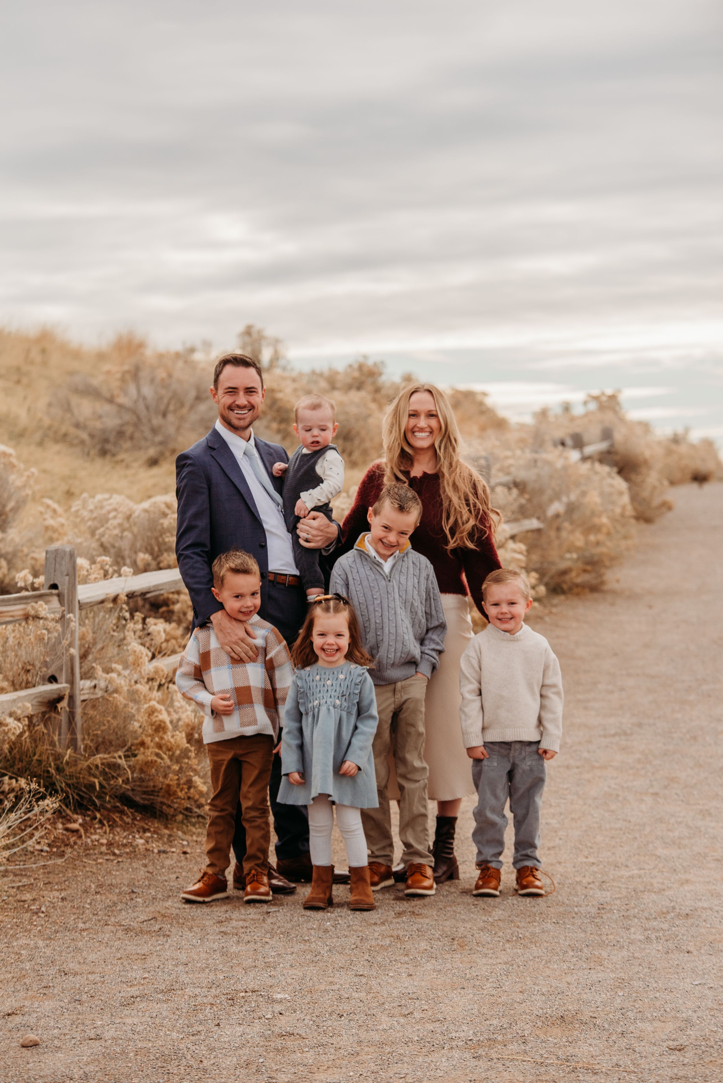 A family of seven poses together on a desert path surrounded by dry brush on an overcast day.