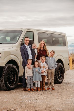 A family stands together in front of a large beige van on a dirt road with cloudy skies above.