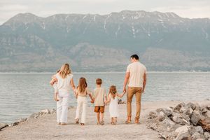 A family in white outfits holds hands while standing on a rocky shore overlooking a mountain lake at sunset.