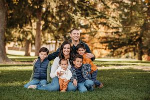 A family group sits together on green grass during a warm autumn day in a park setting.