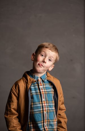 A young child wearing a brown jacket and plaid button-up shirt poses playfully against a gray background.
