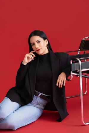 Fashion model in black blazer and jeans poses against red background with modern chair.