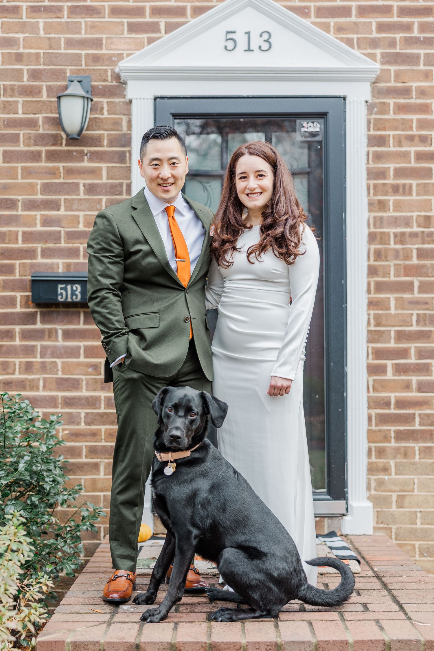 A couple in wedding attire poses with their black Labrador dog on a brick porch.