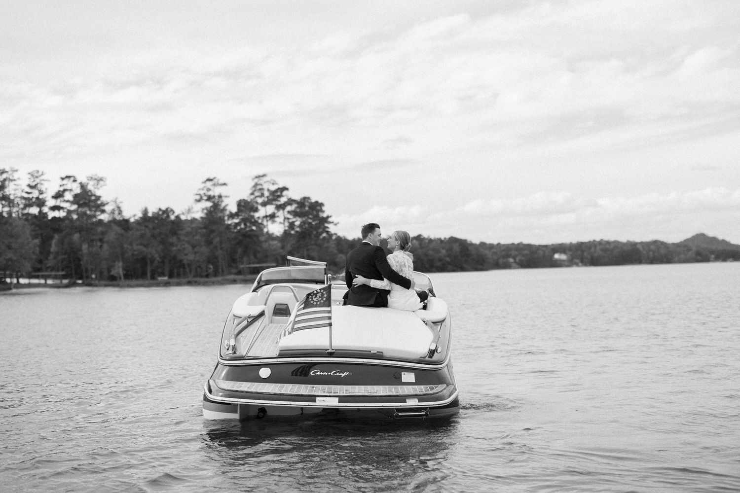 A vintage boat speeds across a calm lake on a sunny day, creating a wake behind it.